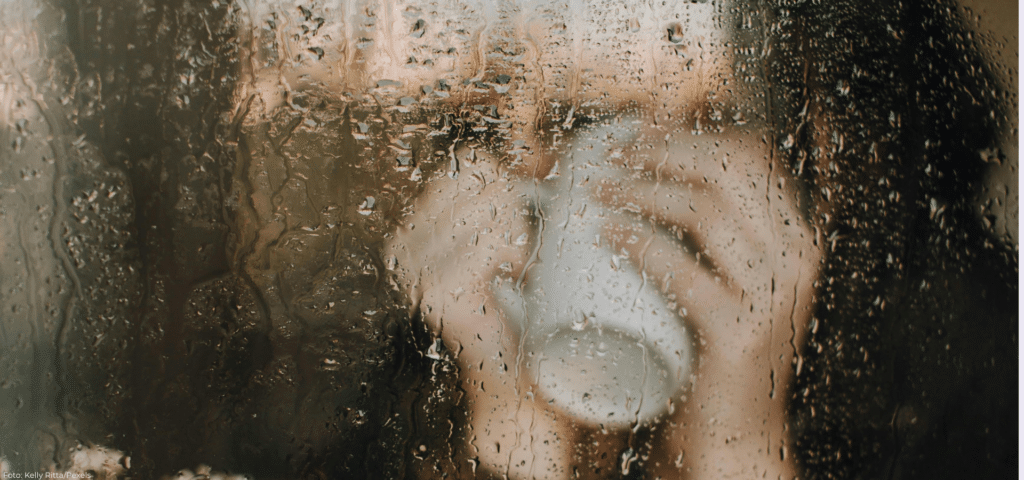 Person hinter einem verregneten Fenster – Symbolbild für Einsamkeit und das Gefühl, durch gesellschaftliche Strukturen unsichtbar zu sein