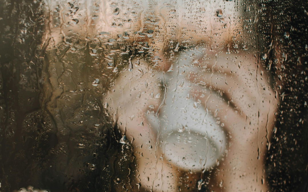 Person mit Kaffeetasse hinter Glasscheibe. Regentropfen trüben die Sicht.