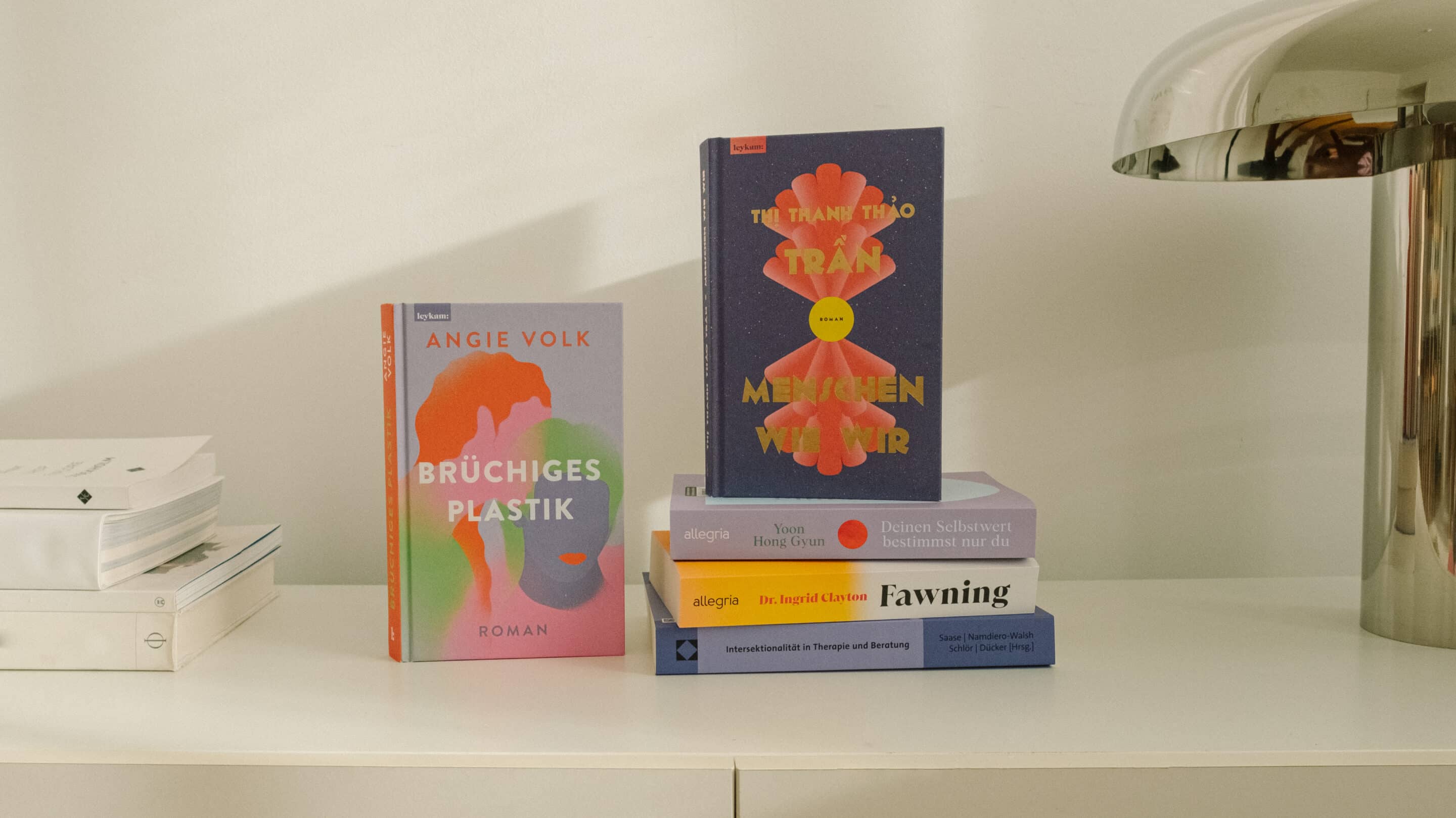 Fünf Buchempfehlungen des ZIMT Magazins auf einem weißen Regal: „Brüchiges Plastik" von Angie Volk, „Menschen wie wir" von Thị Thanh Thảo Trần, „Deinen Selbstwert bestimmst nur du" von Yoon Hong Gyun, „Fawning" von Dr. Ingrid Clayton und „Intersektionalität in Therapie und Beratung" von Saase et al.