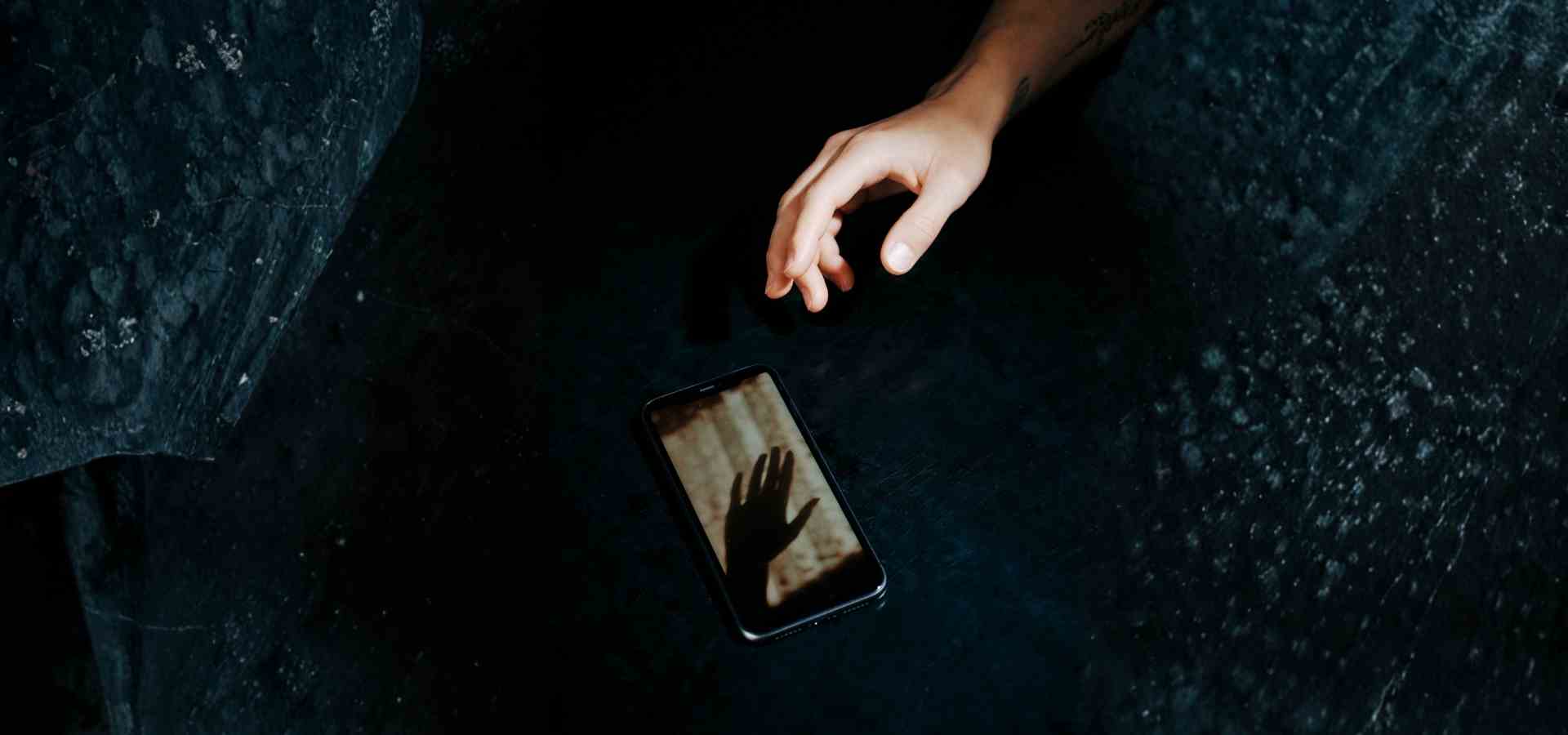 Hand greift nach Smartphone – KI-Chatbot als psychische Unterstützung bei Depressionen Hand greift im Dunkeln nach einem Smartphone, auf dessen Display ein Handschatten zu sehen ist – Symbolbild für die nächtliche Nutzung von KI-Chatbots als psychische Unterstützung