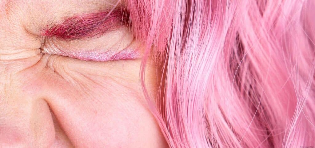 Extreme Nahaufnahme eines fest zusammengepressten Auges mit rosafarbenem Lidschatten und pinken Haaren daneben