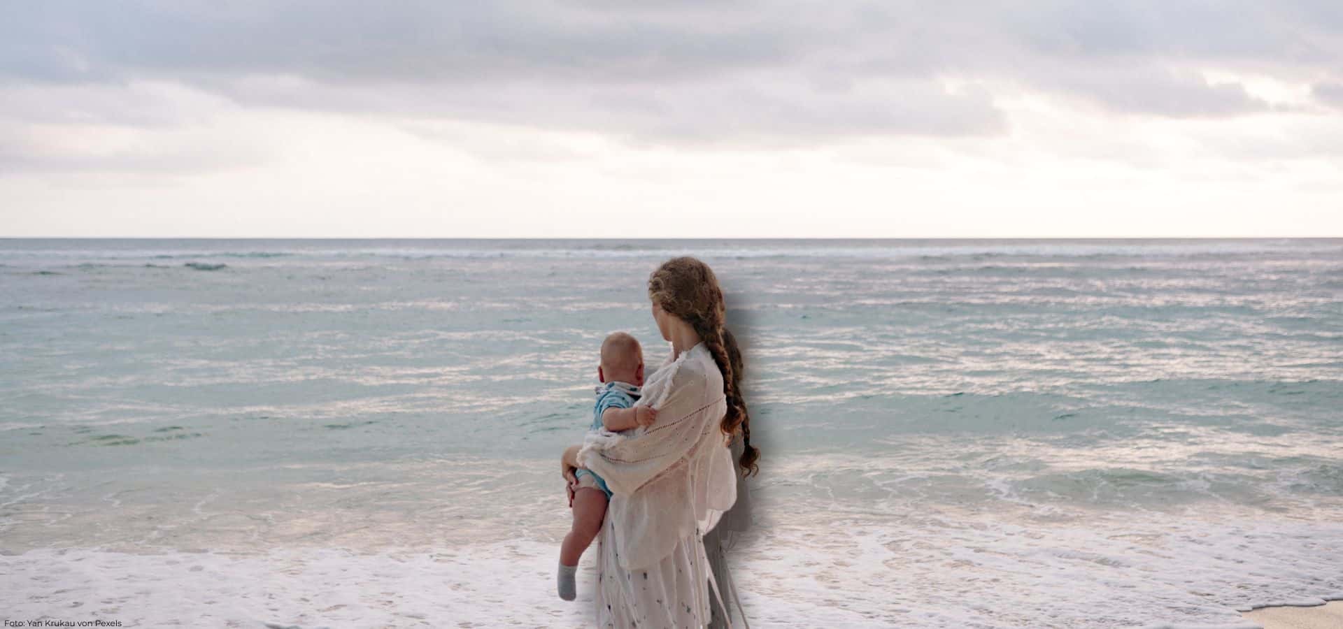 Eine Person mit geflochtenem Haar hält ein Baby im Arm und blickt am Strand auf das Meer hinaus.
