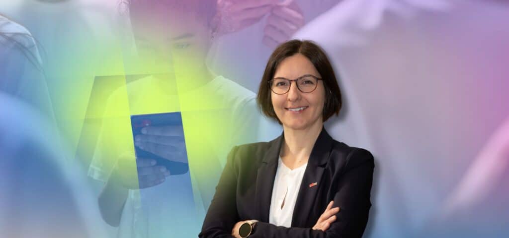 Elisabeth Lex mit Brille und dunklem Blazer, Arme verschränkt, lächelt. Links ein Kind, das auf ein Smartphone schaut.