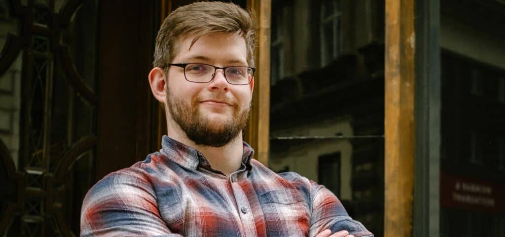 Marcus, junger Mann mit Brille und Bart, im karierten Hemd mit verschränkten Armen vor einem Altbau-Eingang, lächelt leicht.